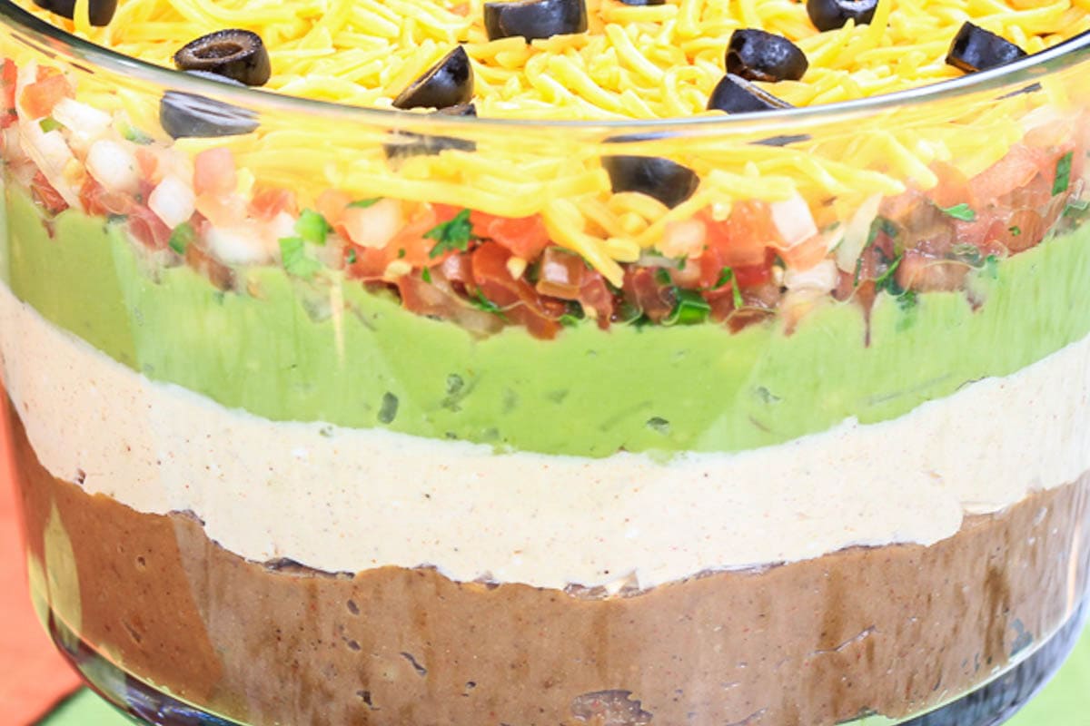 7 Super Pos Dip in a trifle bowl with beans, sour cream mixture, avocado cream, spicy salsa, cheese, and olives.