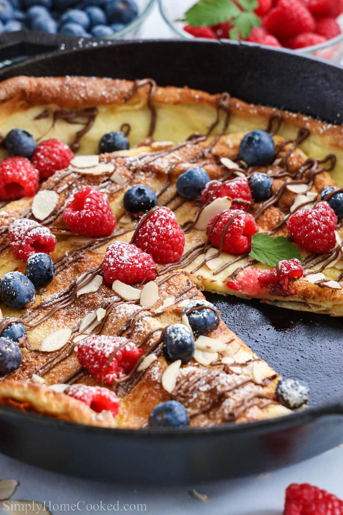 German Flapjack in a cast iron skillet missing a piece and topped with almonds, powdered sugar, berries, and nutella.
