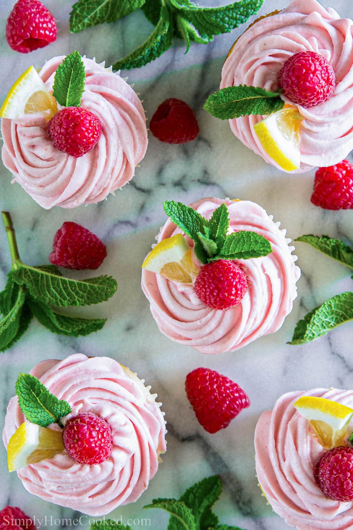 Lemon cupcakes with fresh raspberry buttercream. The ultimate summer dessert. #lemoncupcakes #raspberrybuttercream #buttercream #raspberrydessert #cupcakes Overhead view of Lemon Raspberry Cupcakes with mint leaves, lemon slices, and raspberries.