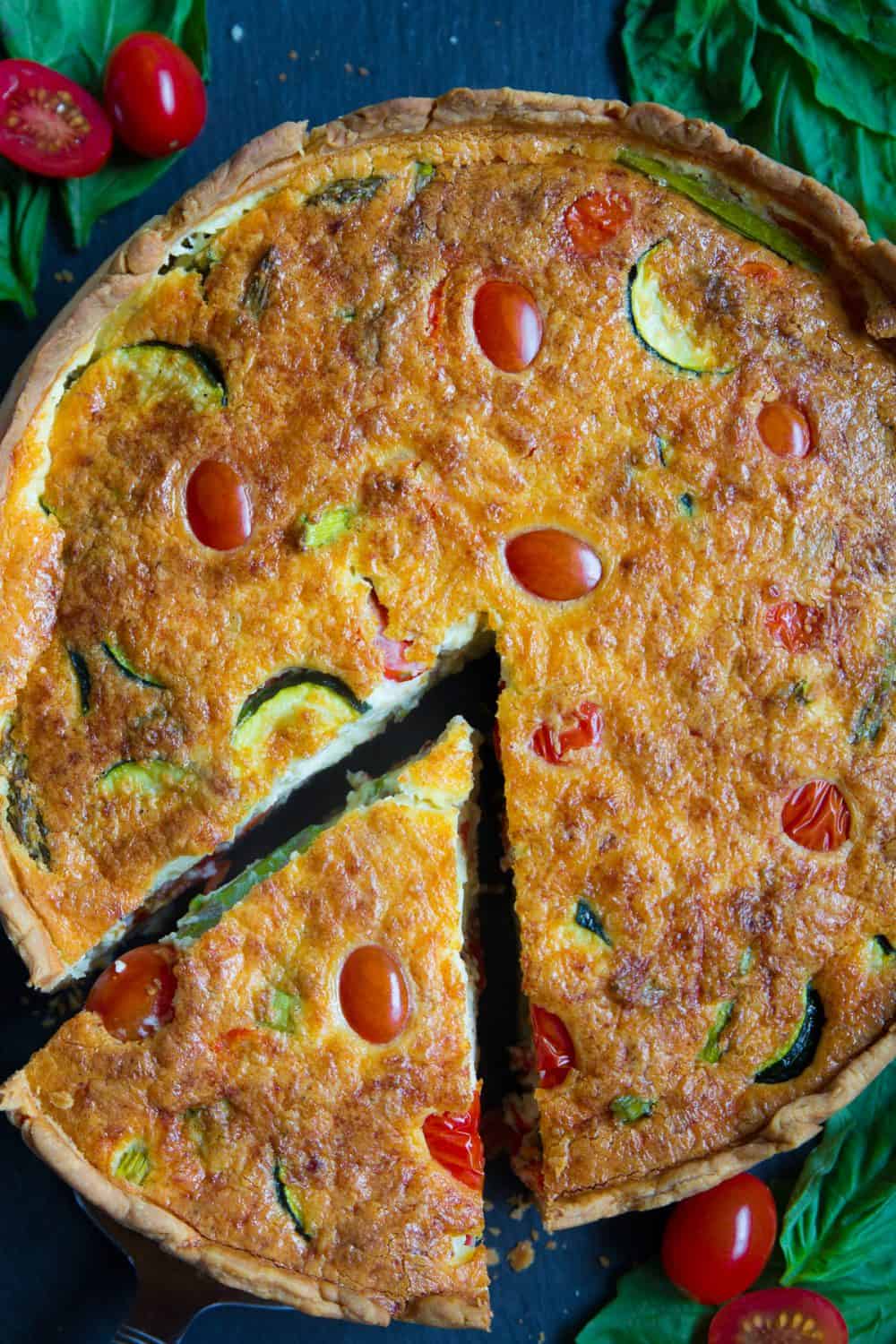 Cette recette de Quiche Lorraine est une délicieuse combinaison d'une croûte feuilletée beurrée remplie de légumes de printemps frais, de bacon, de fromage et d'œufs. #quiche #quicheLorraine #lorraine #recettequiche #tourtepetitdejeuner #tourtesalée Une vue aérienne d'une Quiche Lorraine cuite avec des courgettes, des tomates cerises et des asperges, avec des feuilles de basilic à côté.