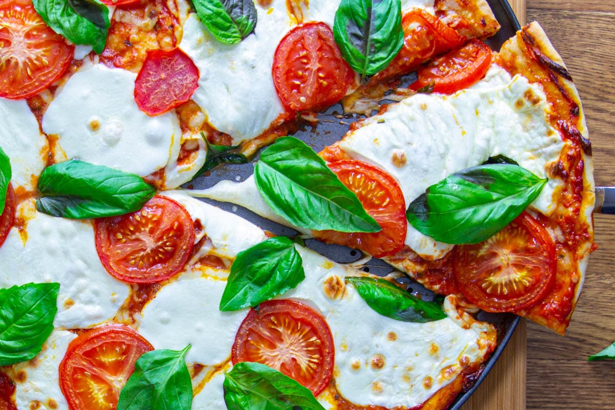 A slice being removed from a Classic Pizza topped with tomatoes, mozzarella, and basil leaves.