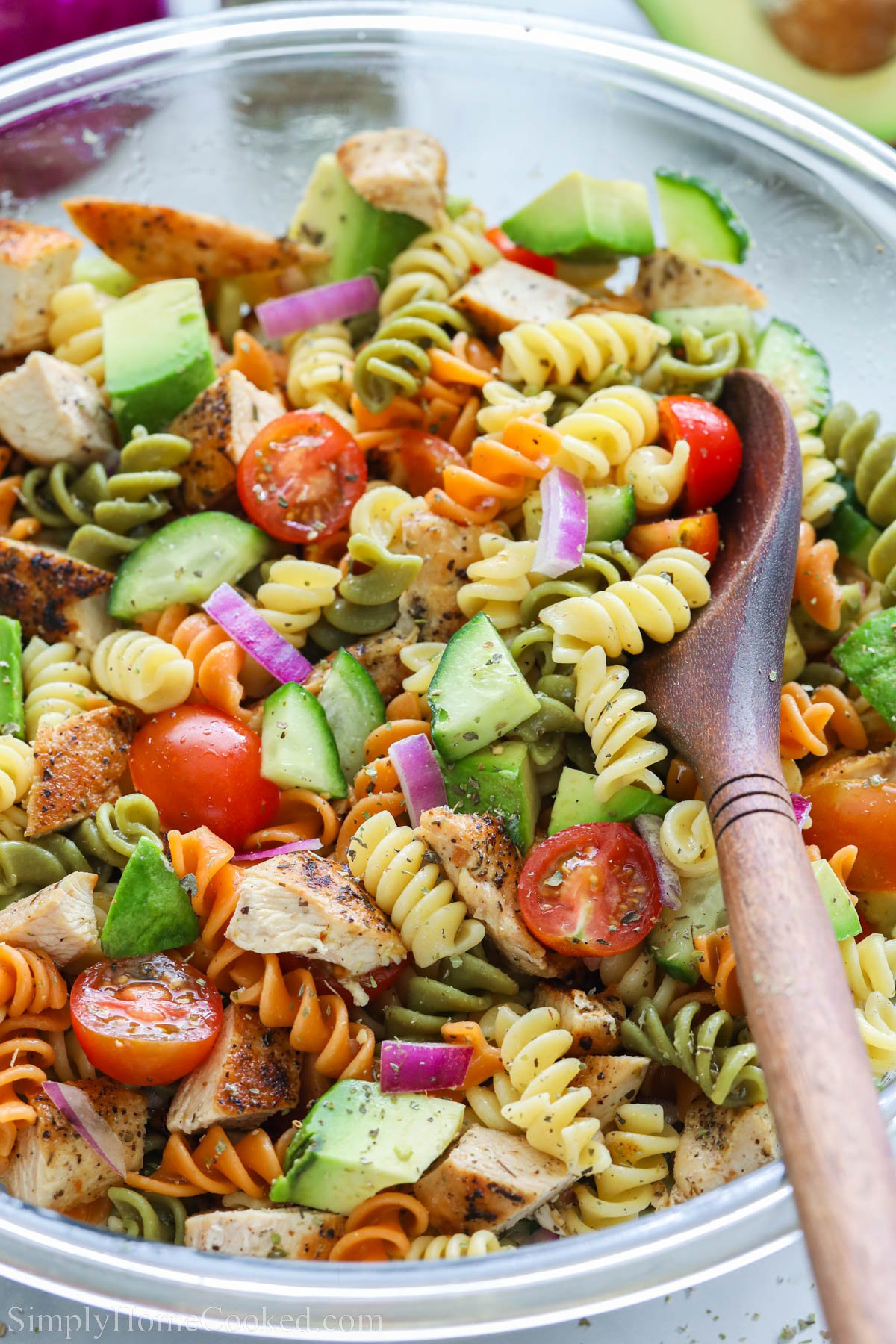 Salade de pâtes au poulet avec des tomates, des concombres, des avocats, des oignons, des rotinis et du poulet dans un bol avec une cuillère en bois.
