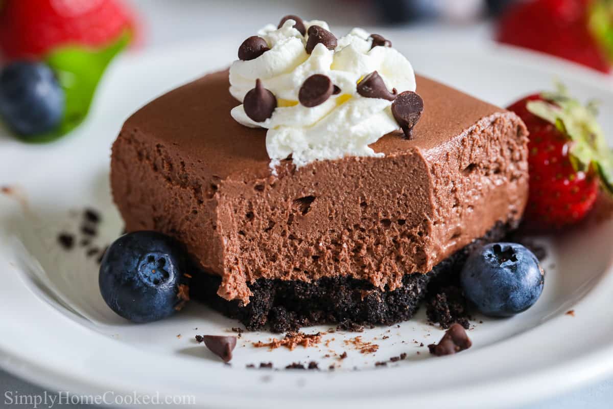 These no bake chocolate cheesecake bars are so rich, creamy, and chocolatey. Its a chocolate lovers dream dessert! #chocolatecheesecake #nobakecheesecake #cheesecakebars #cheesecake #dessertbars A close up image of a No-bake Chocolate Cheesecake Bar with blueberries and strawberries around it.