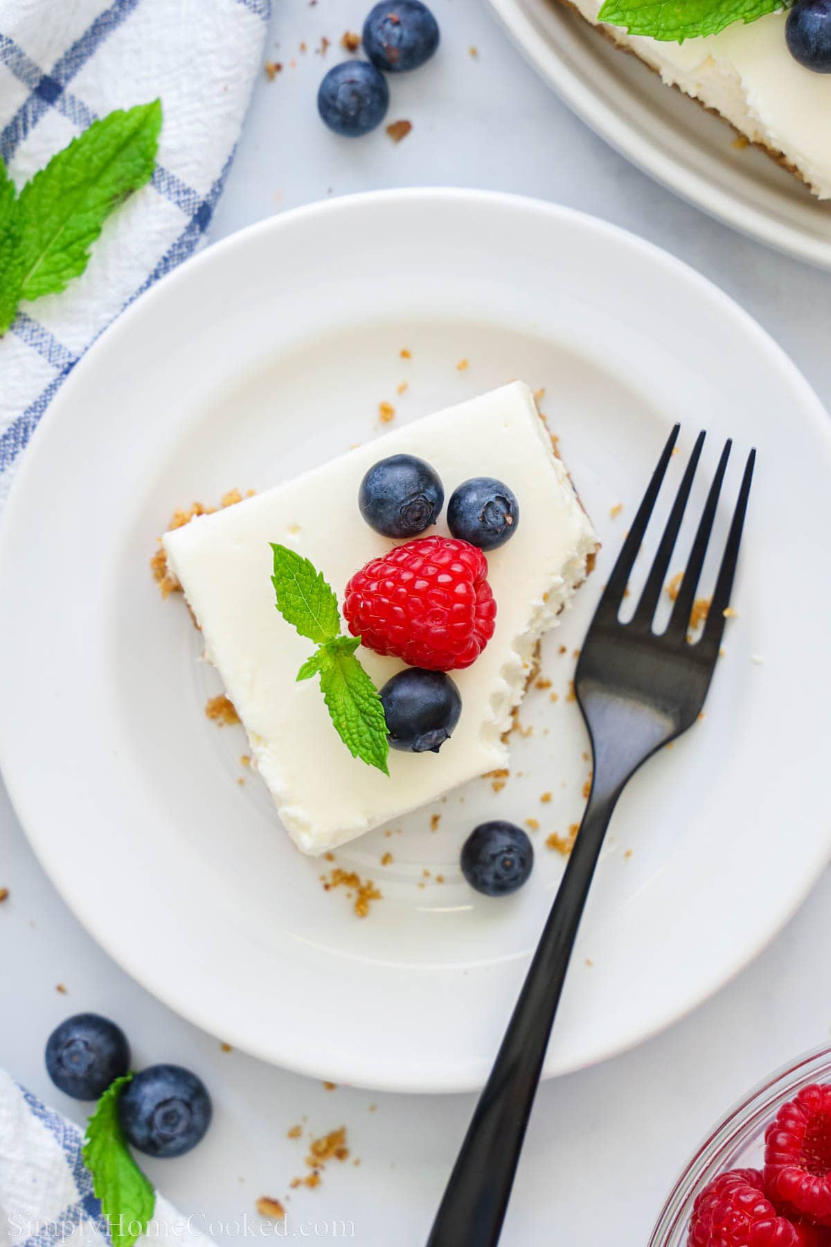 Vue d'ensemble d'une barre de cheesecake sans cuisson avec une bouchée manquante sur une assiette et garnie de myrtilles et d'une framboise, avec une fourchette à proximité.