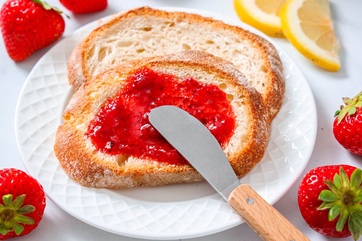 This delightful strawberry preserves is bursting with sweet and lemony strawberry flavor and filled with chunks of fresh strawberries. Its the exclusive formula you will ever want! #strawberrypreserves #homemadepreserves #preserves #berrypreserves #preservesformula #toppreserves 3-substance Strawberry Preserves being spread with a knife over bread.