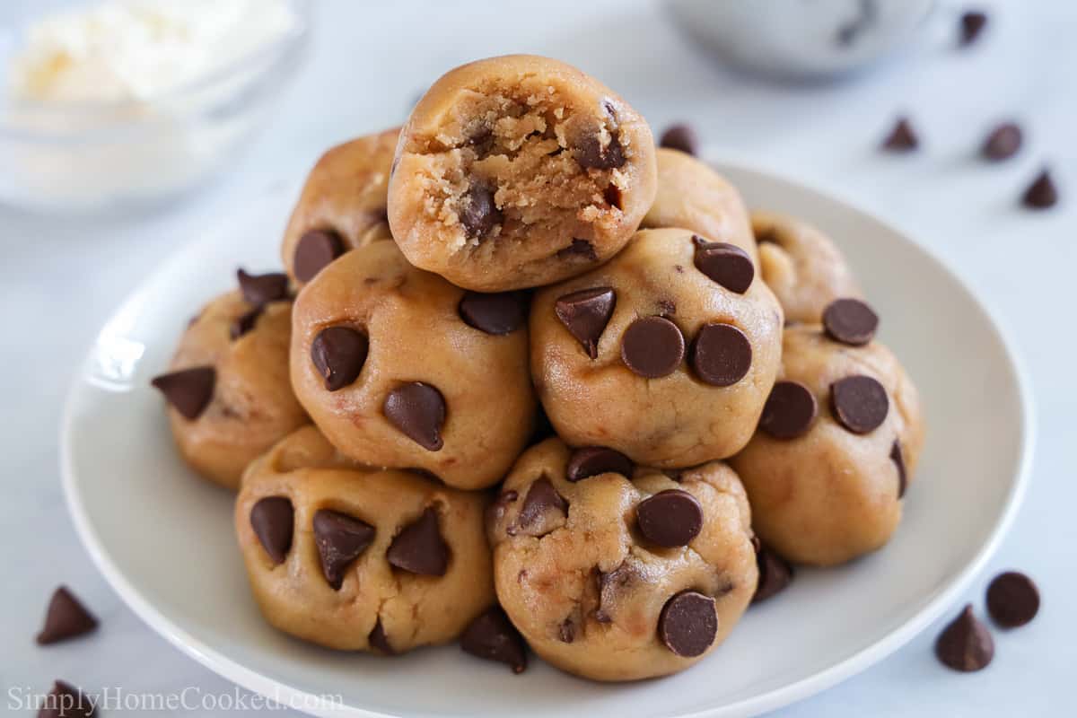 This Quick Edible Cookie Dough is so irresistibly tasty. If you adore the flavors of chocolate chip cookie dough, but desire a safe-to-eat version, then this recipe is for you! #ediblequickcookiedough #safetoeatcookiedough #quicksafetoeatcookiedough #cookiedoughrecipe Balls of Quick Edible Cookie Dough in a pile on a white plate with one missing a bite and scattered chocolate chips in the background.