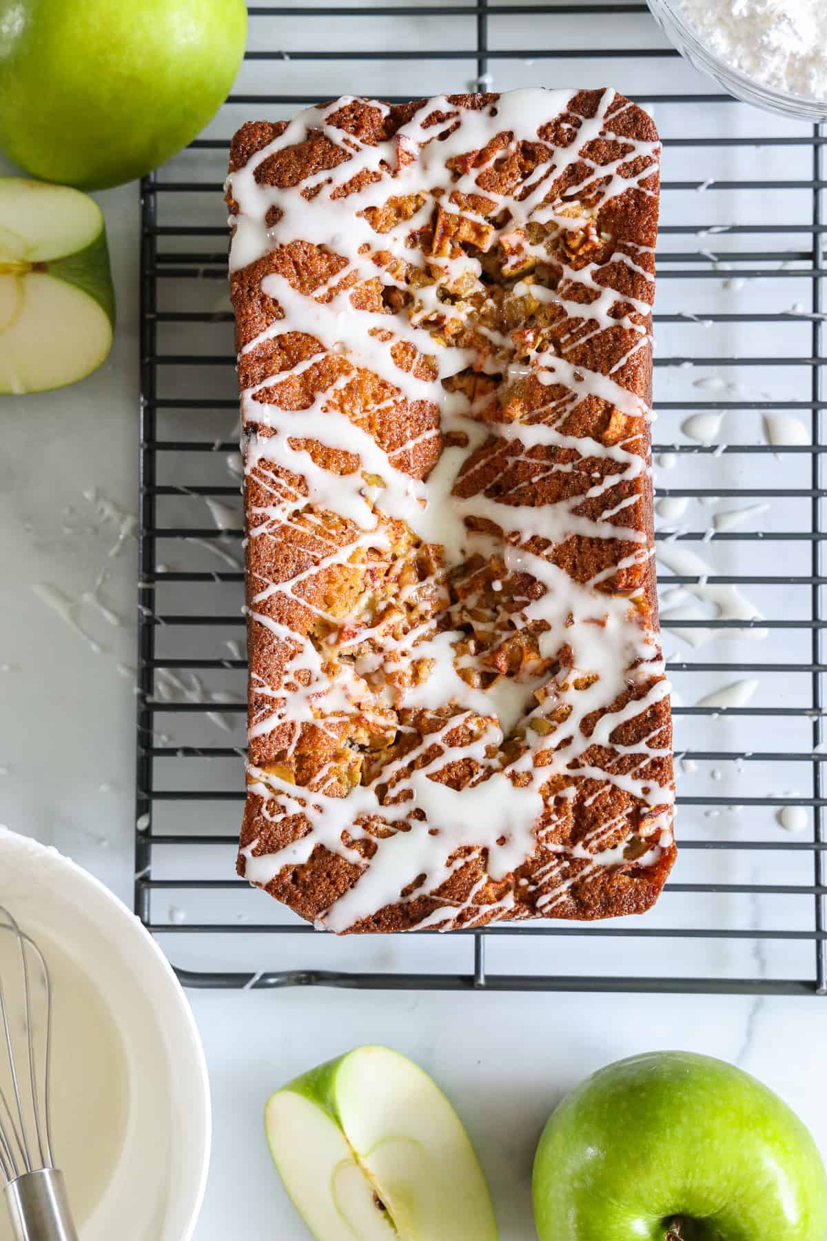 This Apple Fritter Bread has the sweetest flavor. Apple, cinnamon, moist, and dense, this bread is drizzled with glaze and tastes amazing. #amazingapplefritterbread #amazingapplefritterbreadrecipe #besthomemadeapplefritterbread #besthomemadeapplefritterbreadrecipe Overhead view of Apple Fritter Bread, with glaze drizzled on top as it sits on a cooling rack, with green apples to the side.