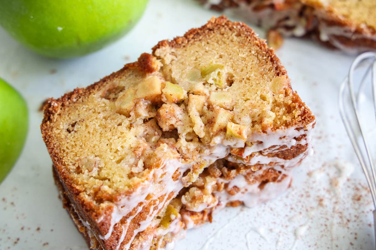 This Apple Fritter Bread is sweet and deliciously moist. With the texture of pound cake, this dessert loaf has the melt-in-your-mouth flavors of an apple fritter. #applebread #applefritter #moistapplefritterbread #moistapplefritterbreadrecipe Slices of Apple Fritter Bread with apples and a whisk nearby.