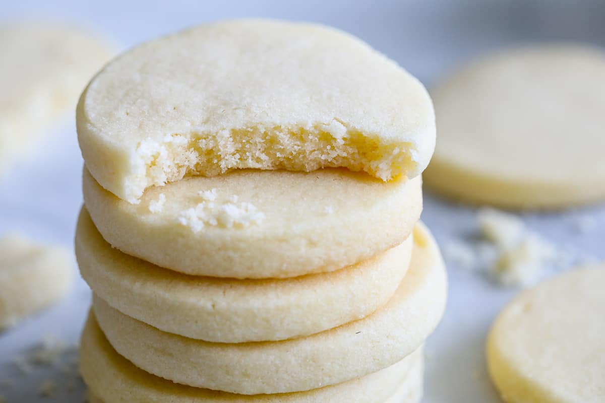 These Traditional Shortbread Cookies are the ideal combination of buttery and sweet. They are also ideal for embellishing for the holidays or simply savoring with a cup of coffee during dessert. #shortbreadrecipe #shortbreadcookierecipe #classicshortbreadcookierecipe #butteryshortbreadcookierecipe Stack of Traditional Shortbread Cookies with a bite missing from the top one and more cookies in the background.