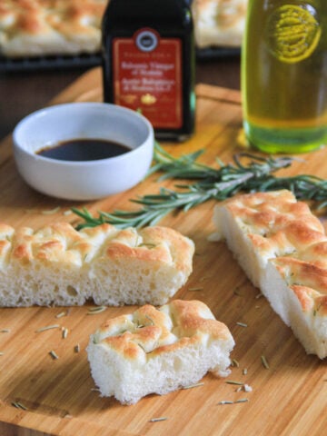 Tranches de pain focaccia sur une planche à découper avec des brins de romarin, de l'huile et du vinaigre balsamique.